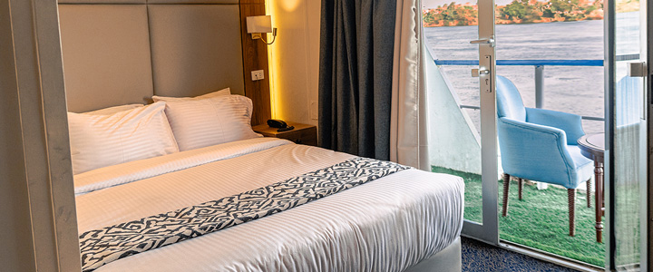 Die moderne Kabine eines Nilkreuzfahrtschiffs ist mit einem Doppelbett und hellen Möbeln eingerichtet. Vom Zimmer führt eine Tür direkt auf den privaten Balkon.