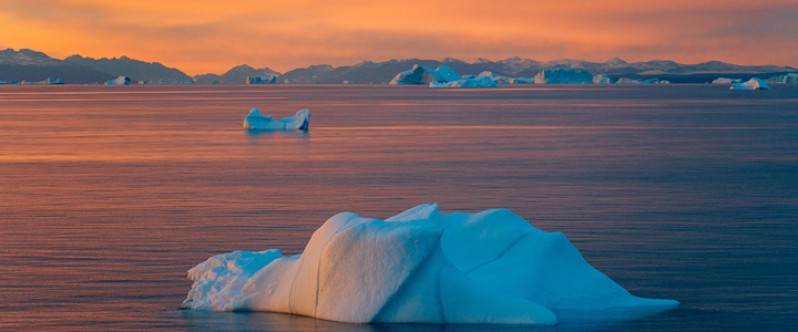 Treibende Eisberge liegen im ruhigen Wasser vor einer dunklen Bergkulisse. Das warme Licht des Sonnenuntergangs taucht Himmel und Meer in sanfte Farben.